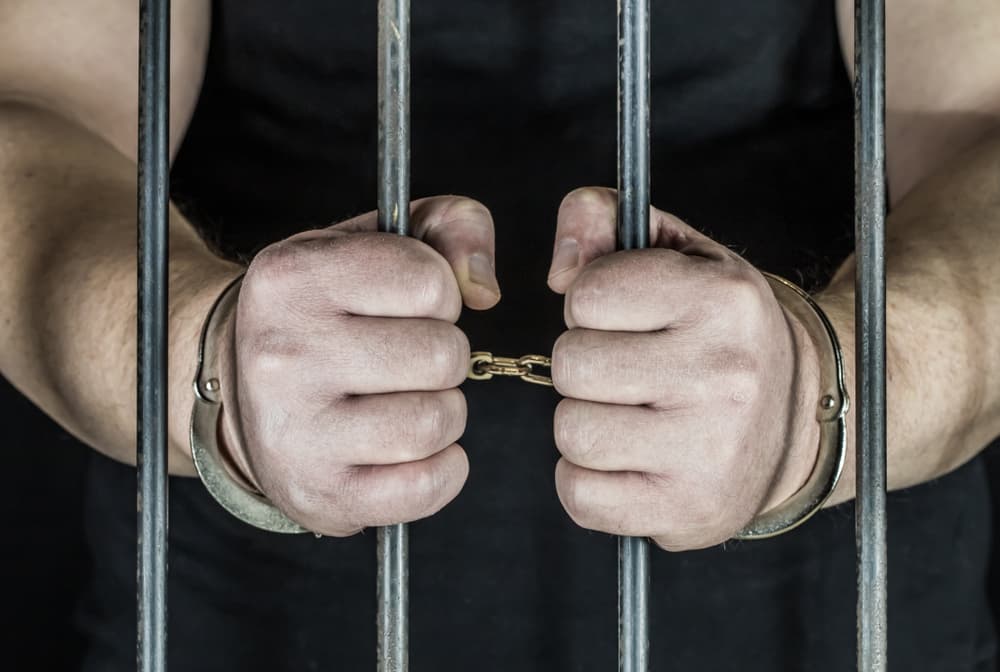 Closeup of a man in handcuffs standing behind prison bars, symbolizing justice and crime.