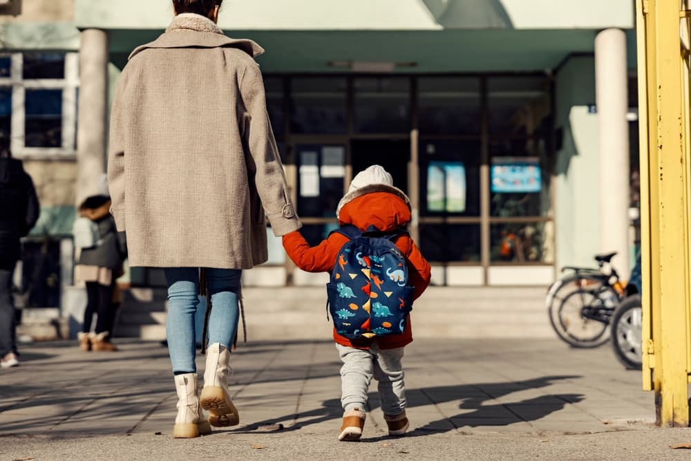 A mother holding hands with her little son and entering the kindergarten yard.